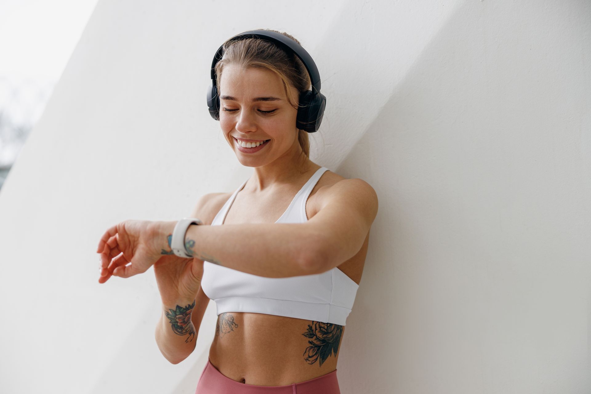 Woman in workout clothes checks smartwatch, smiling, wearing headphones, leaning against a white wall.