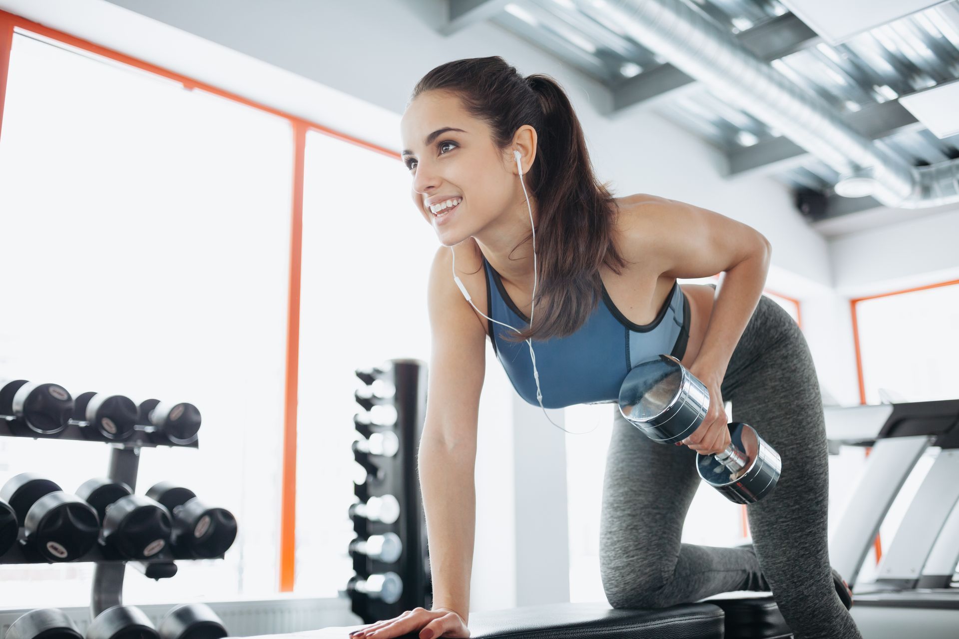 Woman doing dumbbell rows in a gym, wearing a sports bra and leggings, smiling.