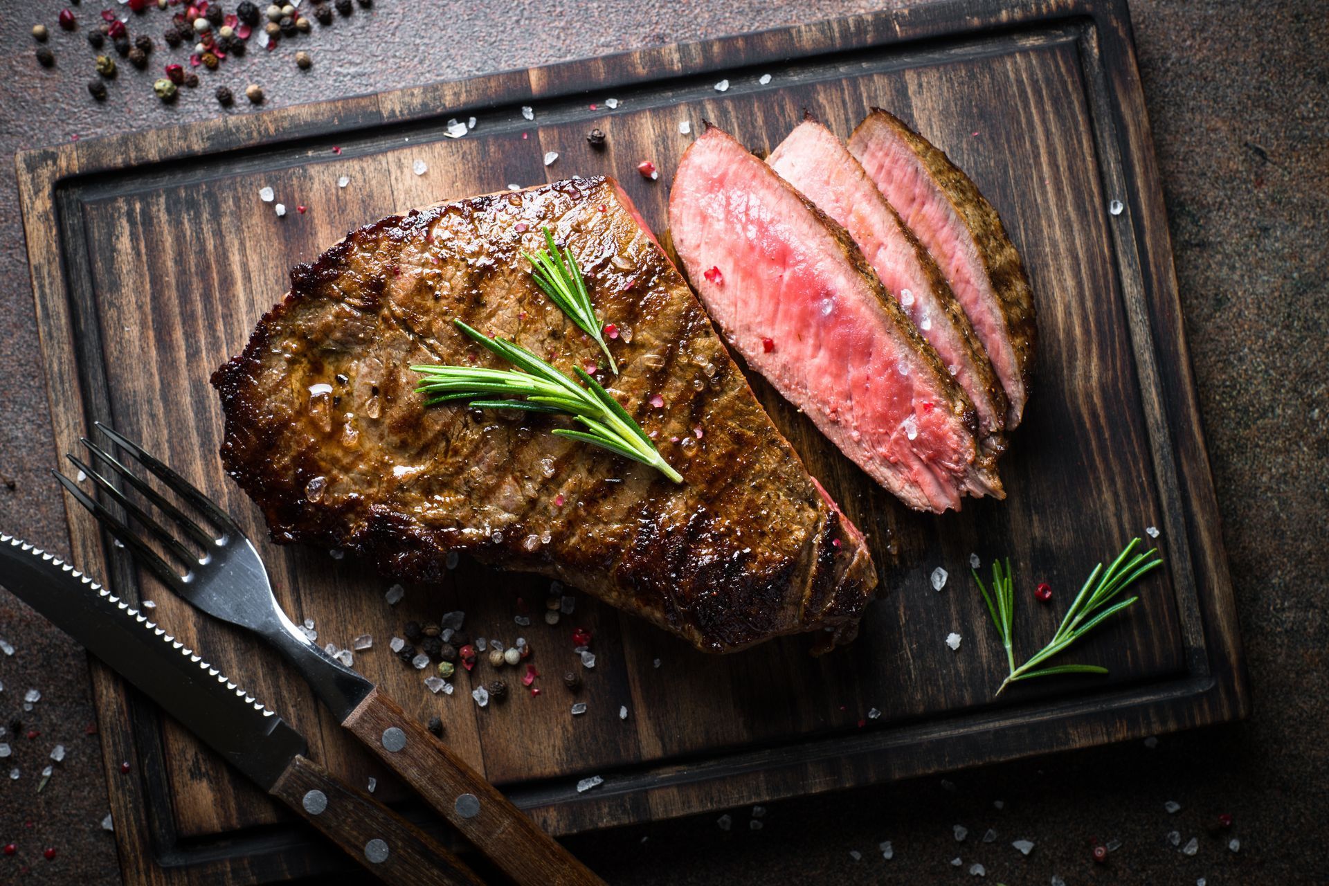 Sliced, grilled steak on a wooden cutting board with a knife, fork, rosemary, and seasoning.