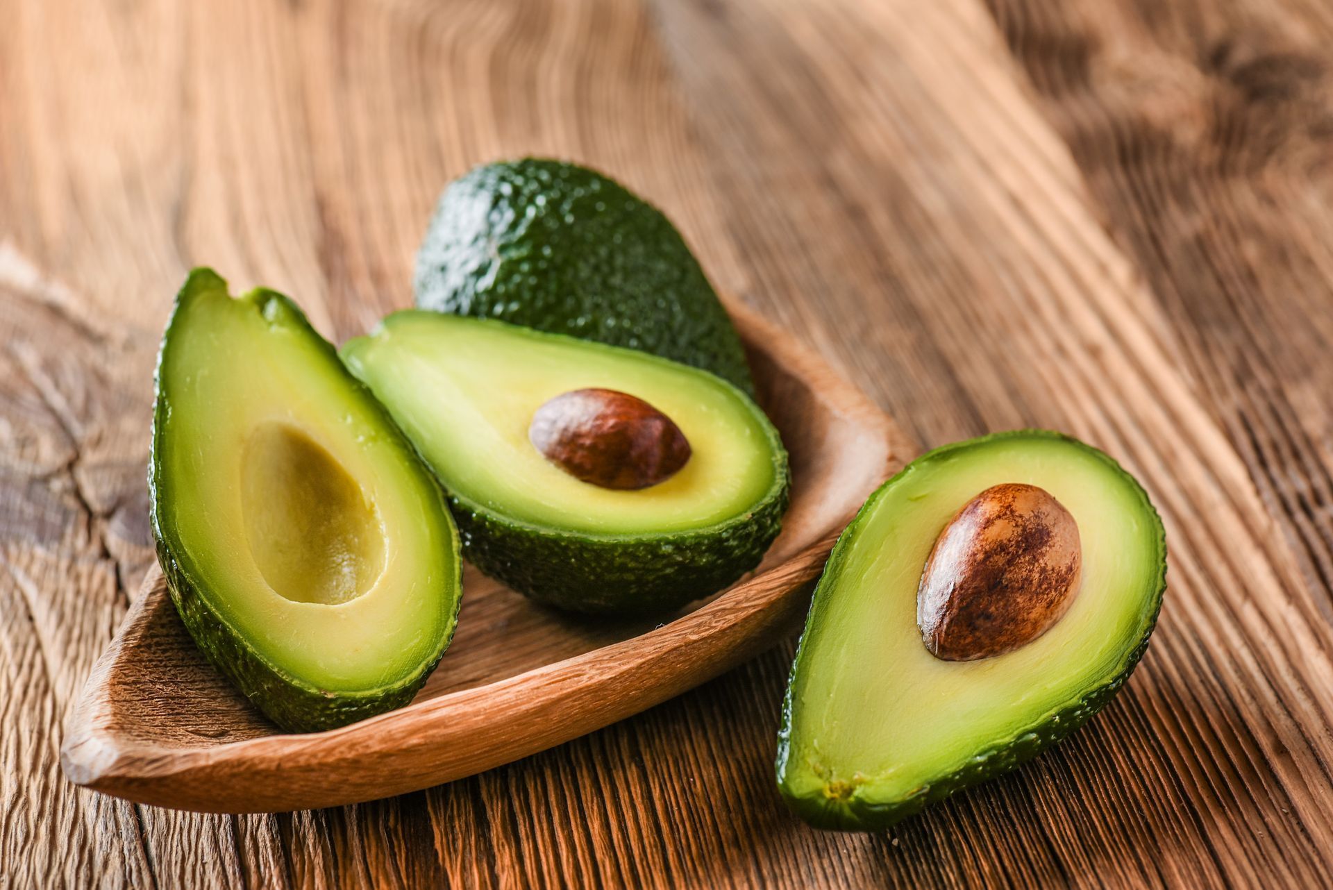 Avocados: ripe green fruit, halved to show creamy insides and brown seed, in wooden bowl on wood surface.