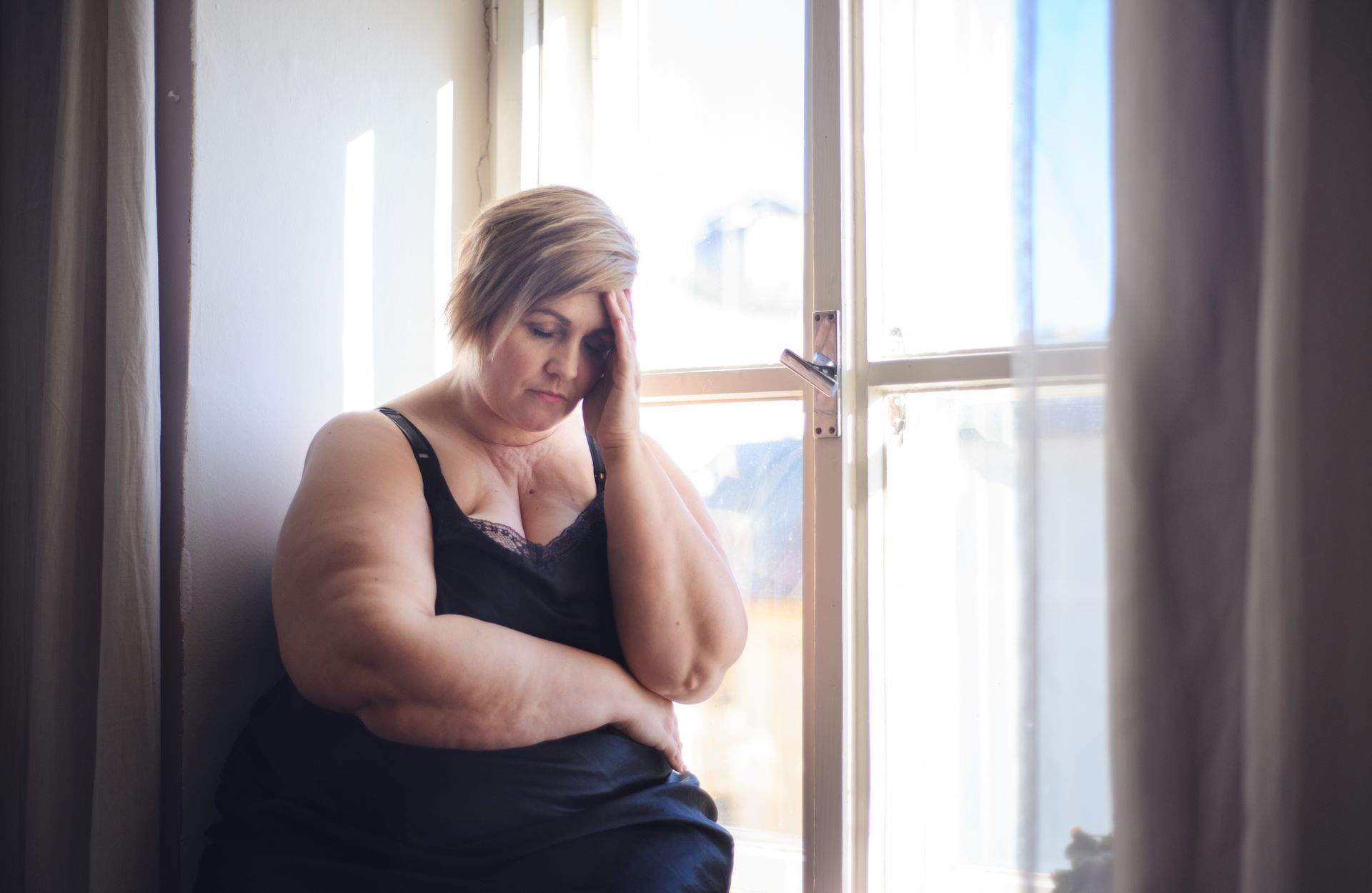 Person in a black dress sits by a bright window, resting their head on one hand, looking down.