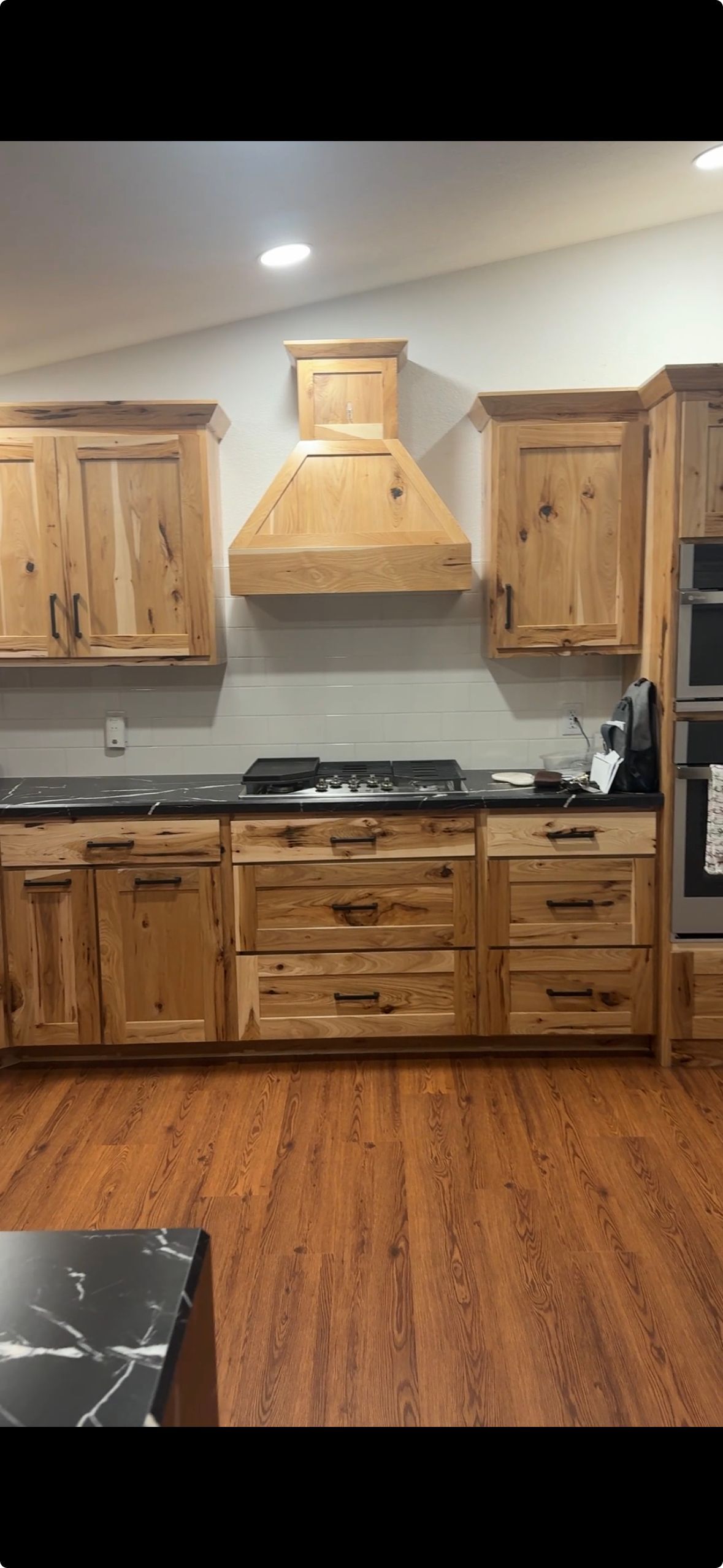 Wooden kitchen with rustic cabinets, black stove, tan range hood, and wood plank flooring.