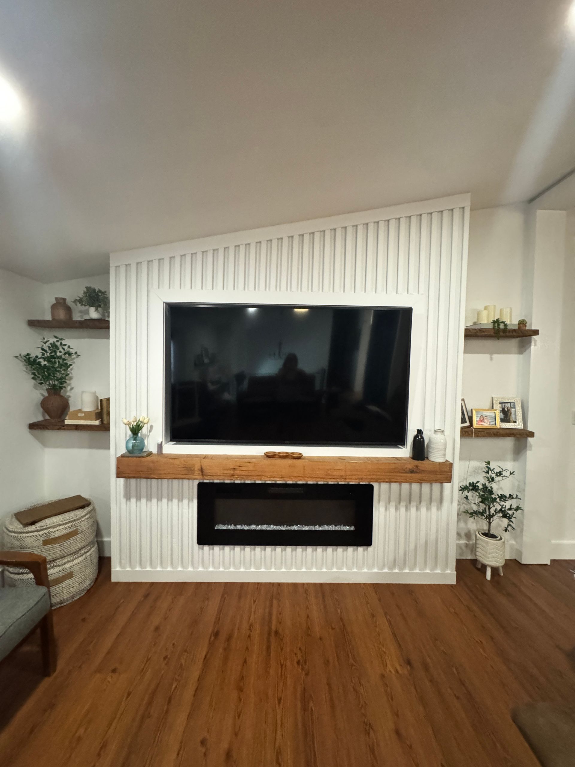 Modern living room with a large TV above a white fireplace and wood floors, flanked by plants and shelves