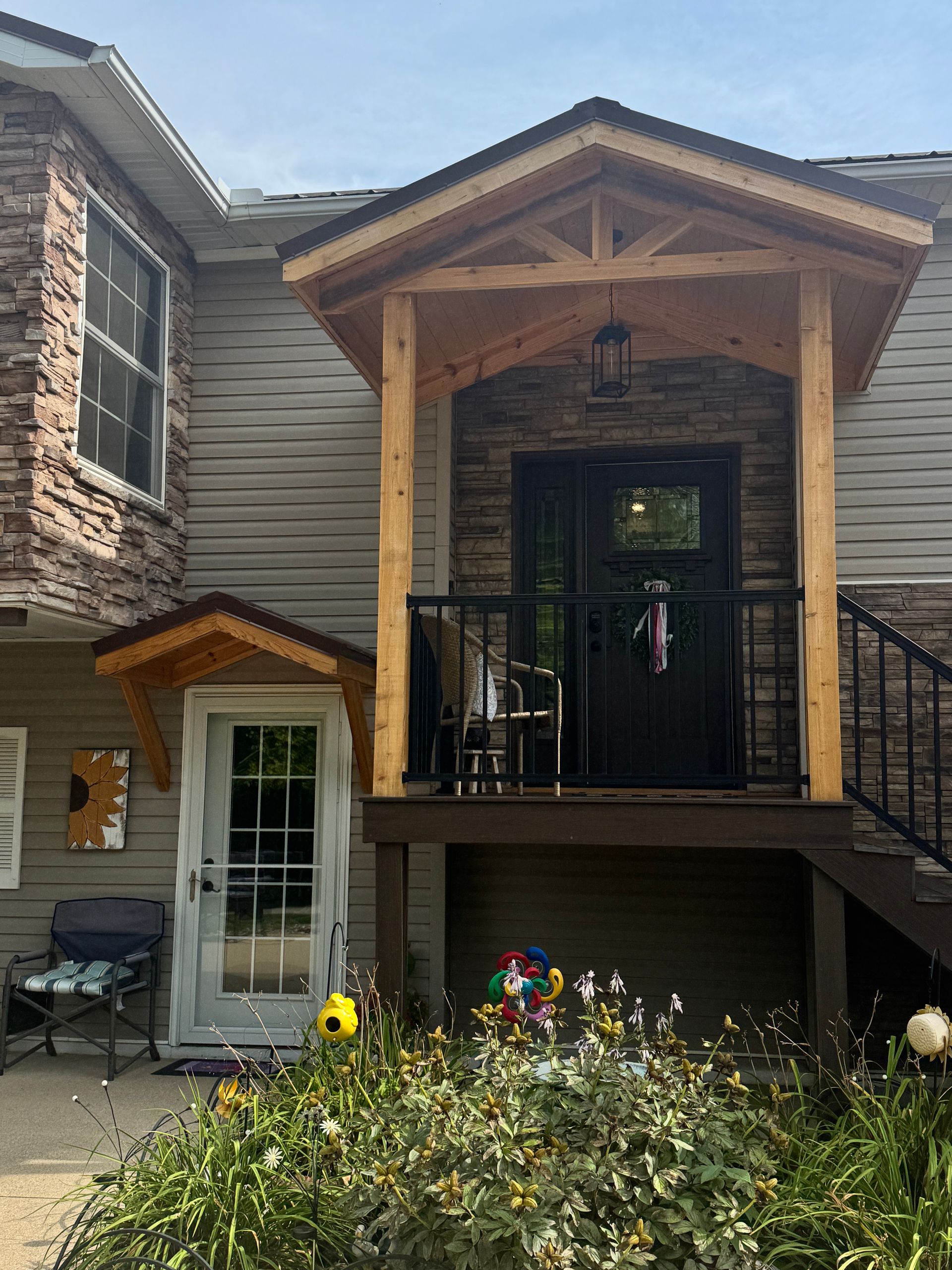 Wood-and-stone house entrance with covered porch, black railing, and flower garden in front