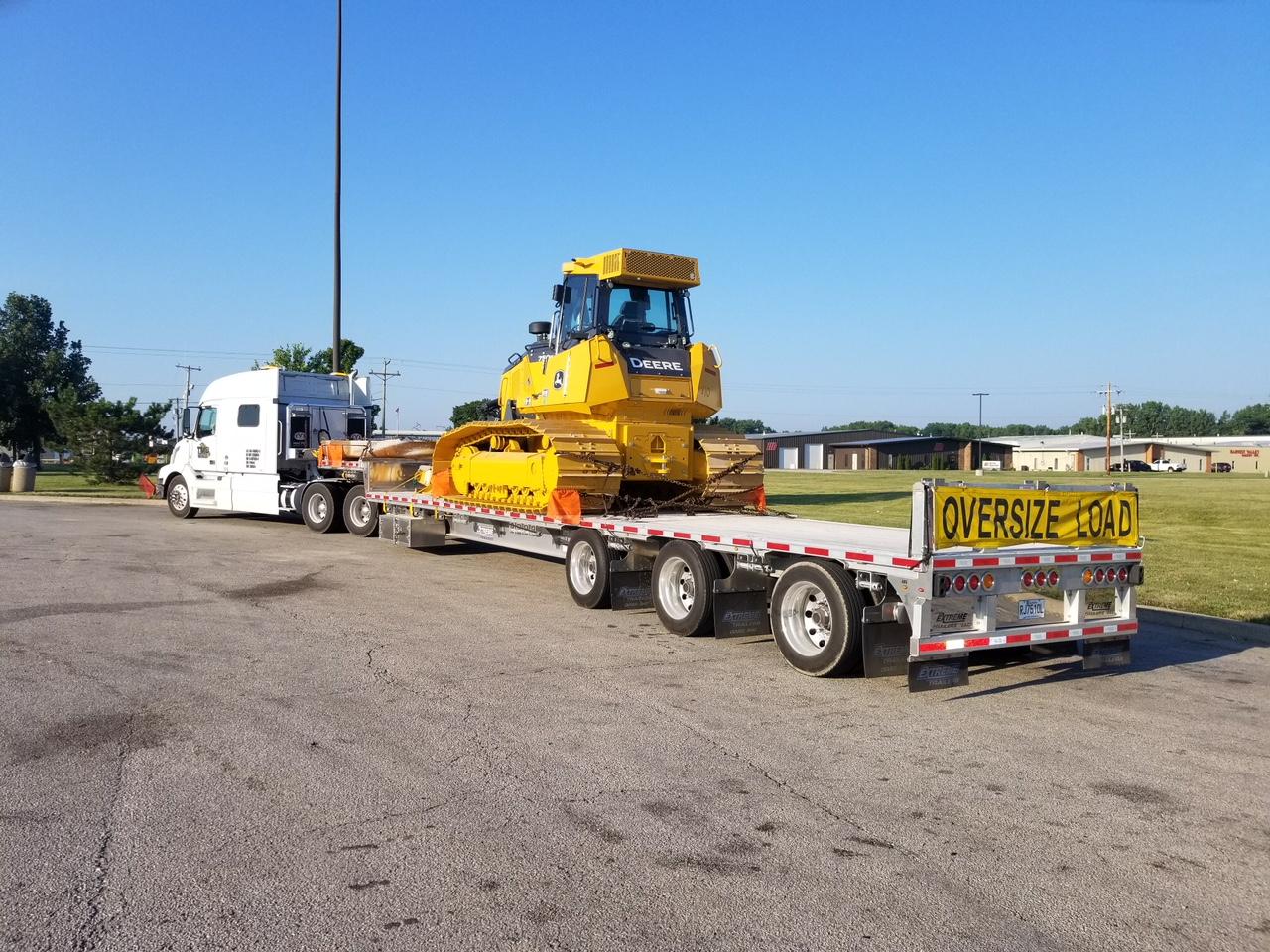 Yellow bulldozer on a flatbed trailer, being hauled by a white truck on a paved lot. 