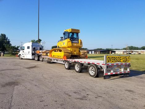 Yellow bulldozer on a flatbed trailer, being hauled by a white truck on a paved lot. 