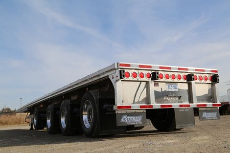 Flatbed trailer, silver with red lights, parked on gravel against a blue sky.