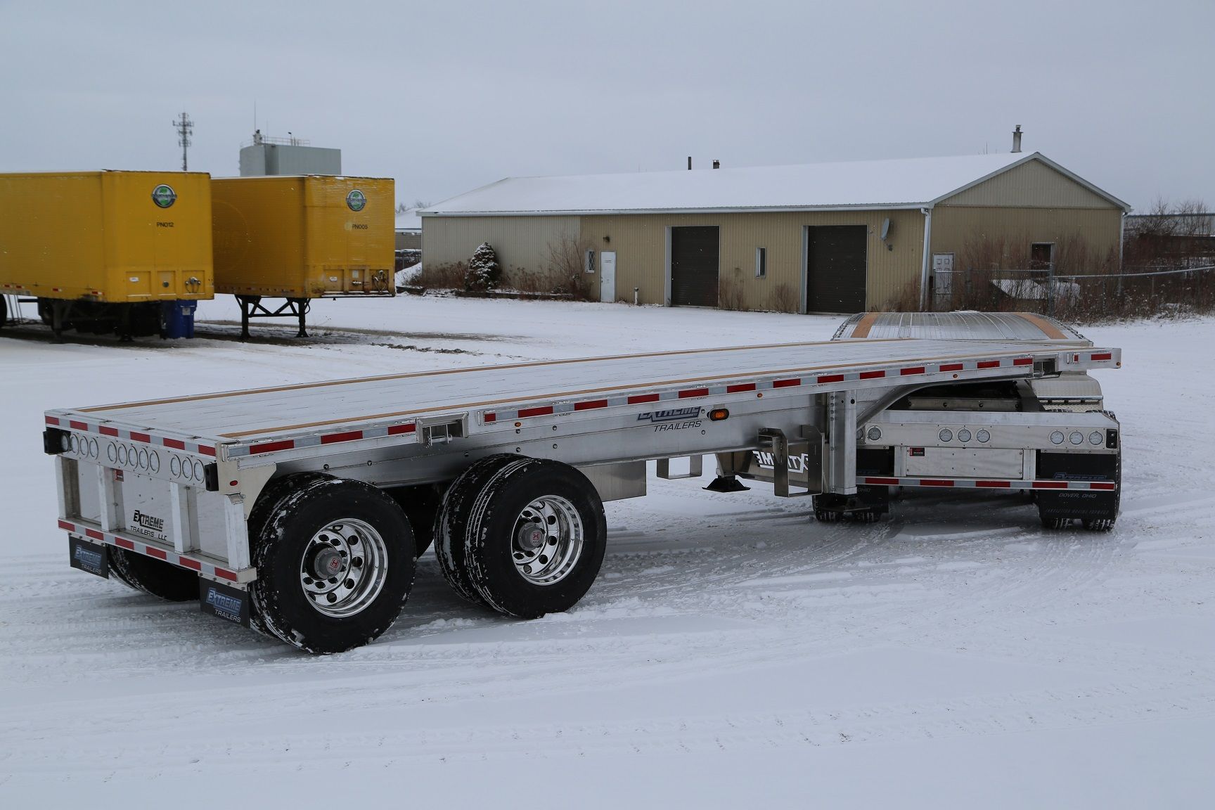Flatbed semi-trailer on snow-covered ground, parked near a building and other trailers on a cloudy day.