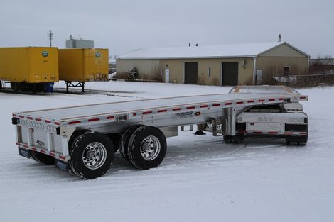 Flatbed semi-trailer on snow-covered ground, parked near a building and other trailers on a cloudy day.