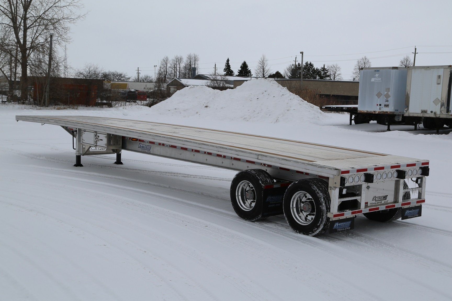 Flatbed trailer on snow-covered ground, parked near buildings and a snow pile.