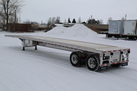 Flatbed trailer on snow-covered ground, parked near buildings and a snow pile.