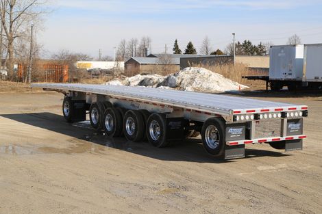 Flatbed trailer on a gravel lot under a clear sky.  The trailer has multiple wheels and a metallic surface.