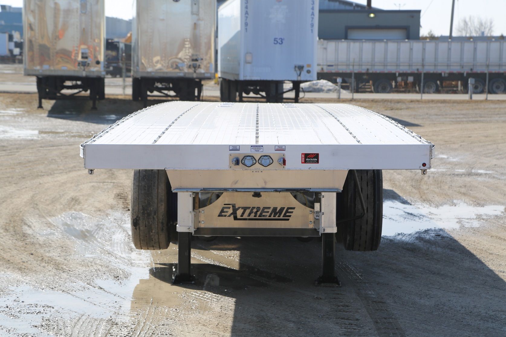 A white flatbed trailer with a black undercarriage is parked outdoors on a sunny day.