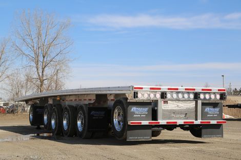 Flatbed trailer on a gravel lot under a blue sky, four axles with chrome wheels, black mud flaps.