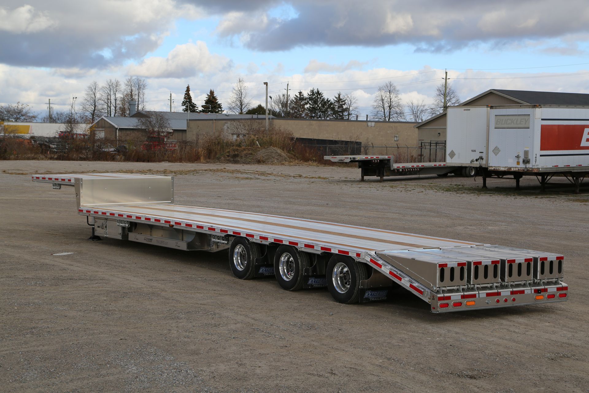Flatbed trailer on gravel lot, with a cloudy sky background and buildings.