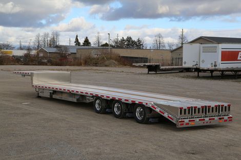 Flatbed trailer on gravel lot, with a cloudy sky background and buildings.