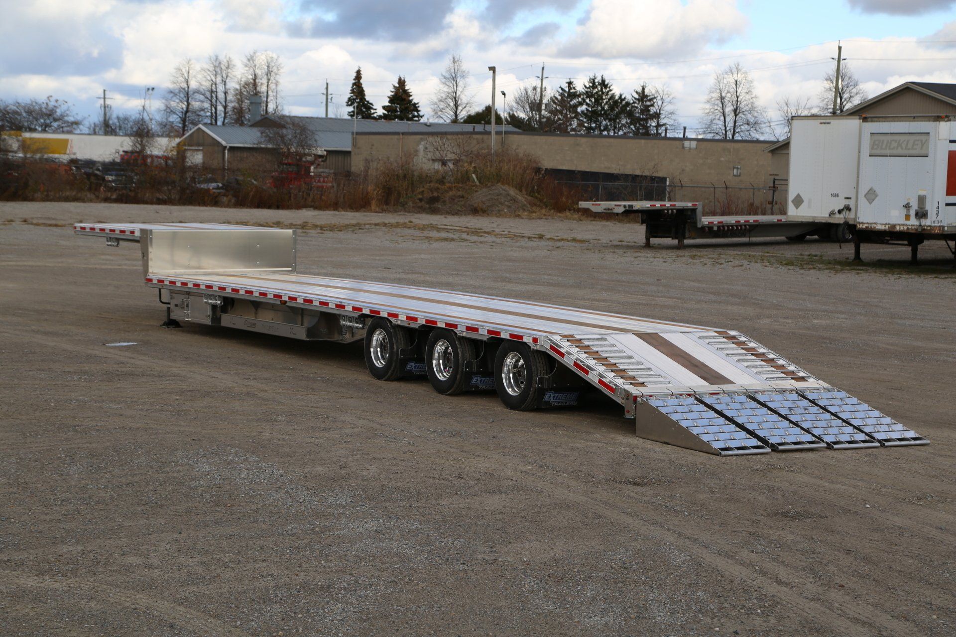 Flatbed trailer with ramps parked on gravel, in an industrial area.