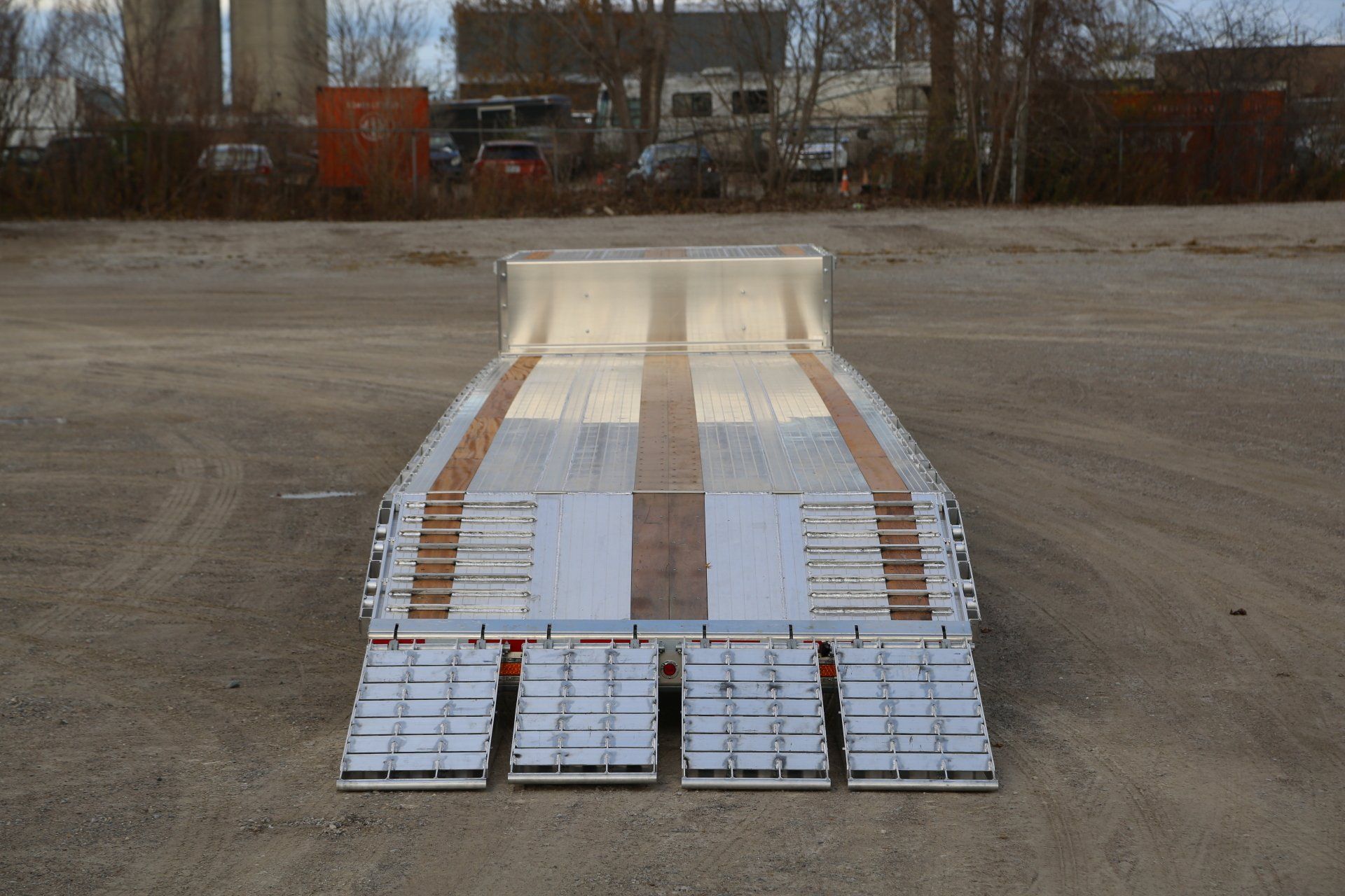 A lowboy trailer with ramps, seen from the front. The trailer bed is silver with wooden inserts, set on a gravel lot.