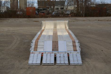 A lowboy trailer with ramps, seen from the front. The trailer bed is silver with wooden inserts, set on a gravel lot.