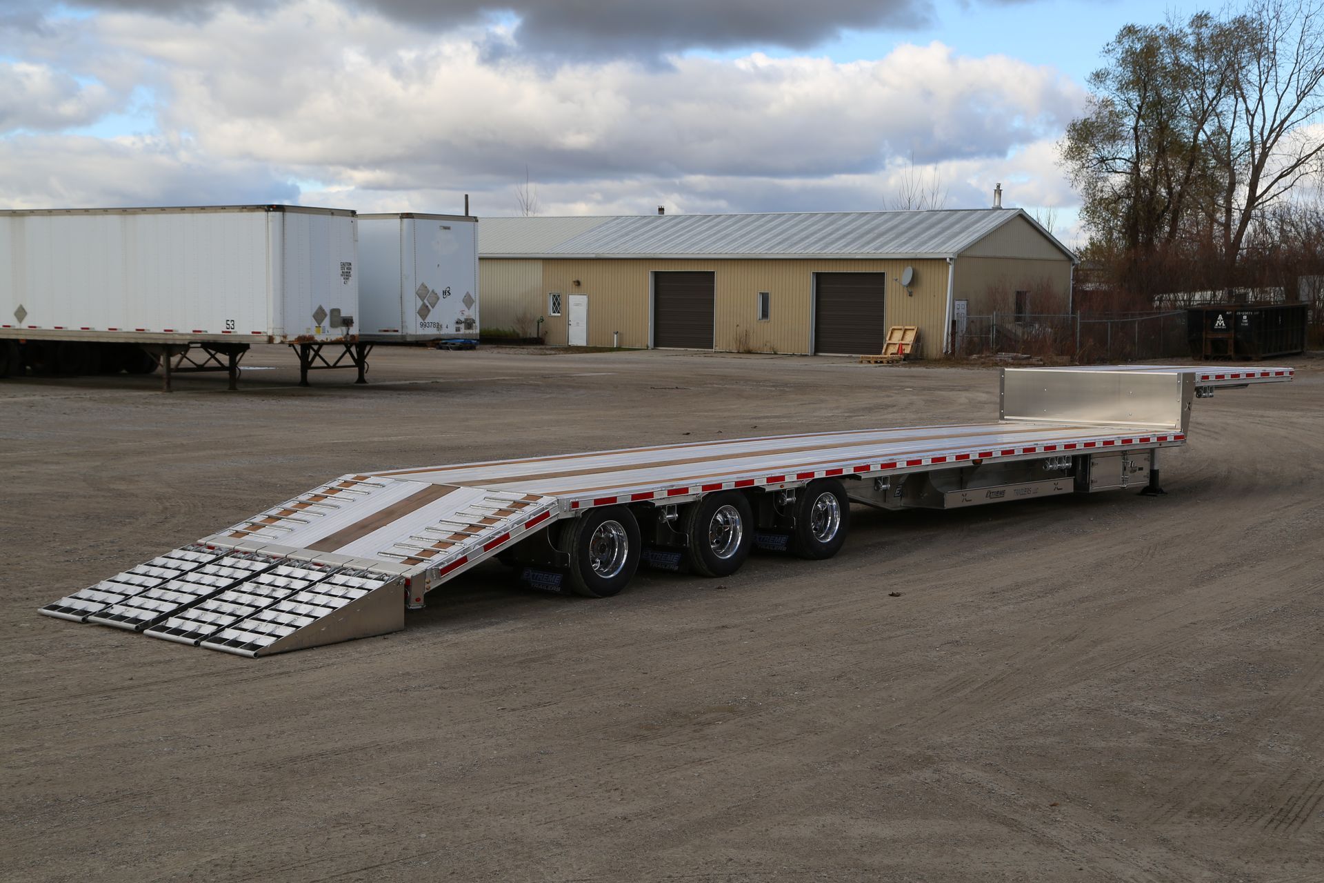 Flatbed trailer with ramps parked on gravel, with warehouses in the background under a cloudy sky.