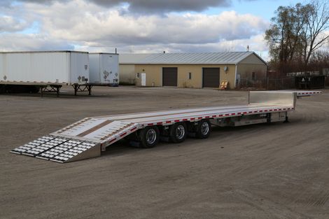 Flatbed trailer with ramps parked on gravel, with warehouses in the background under a cloudy sky.