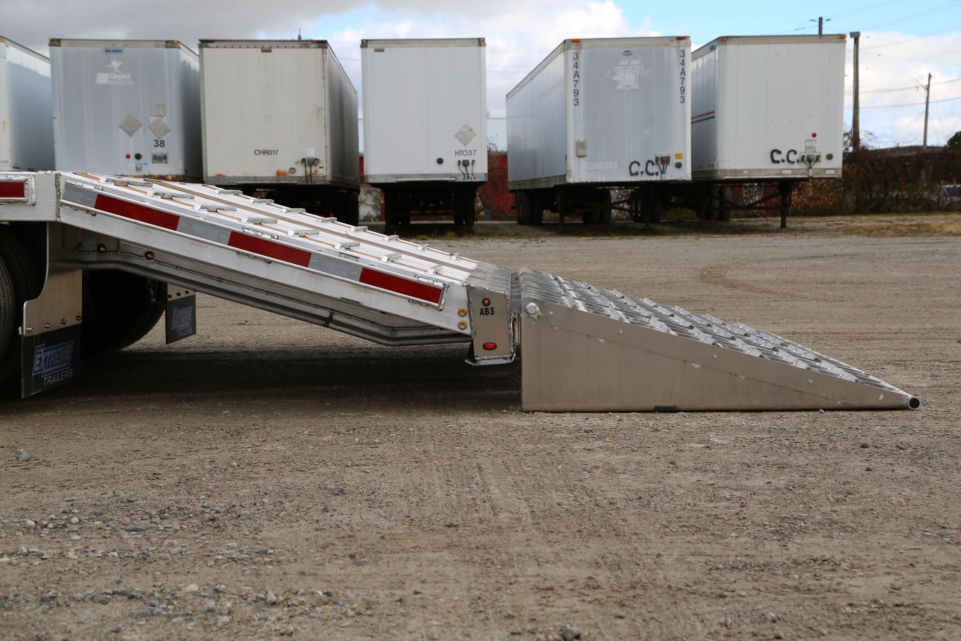 A semi-trailer with an extended ramp for loading or unloading goods. Trailers are parked on gravel.