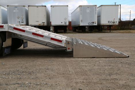 A semi-trailer with an extended ramp for loading or unloading goods. Trailers are parked on gravel.