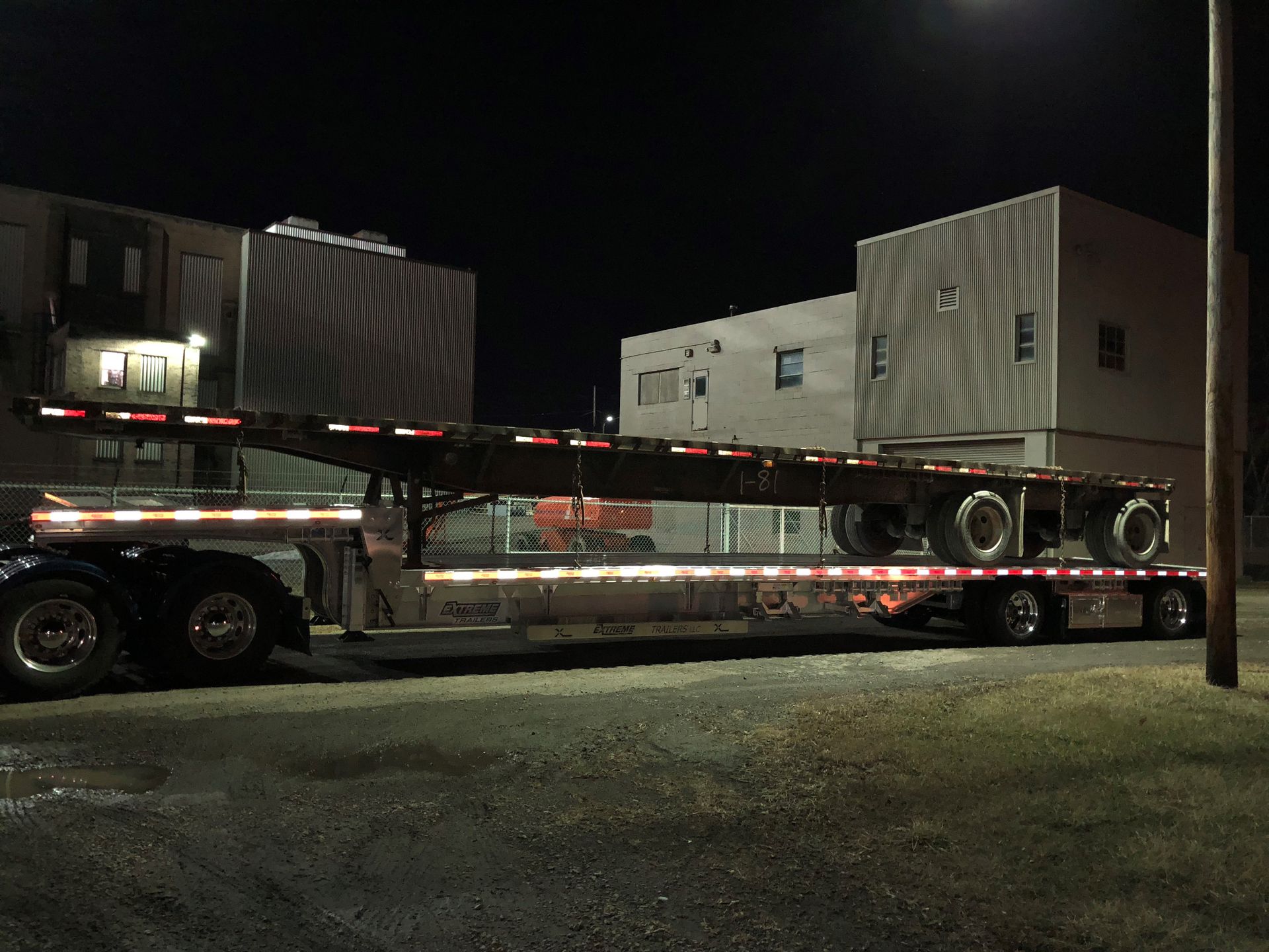 Semi-truck at night hauling a flatbed trailer with reflective tape, parked near buildings.