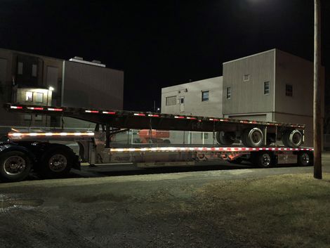 Semi-truck at night hauling a flatbed trailer with reflective tape, parked near buildings.