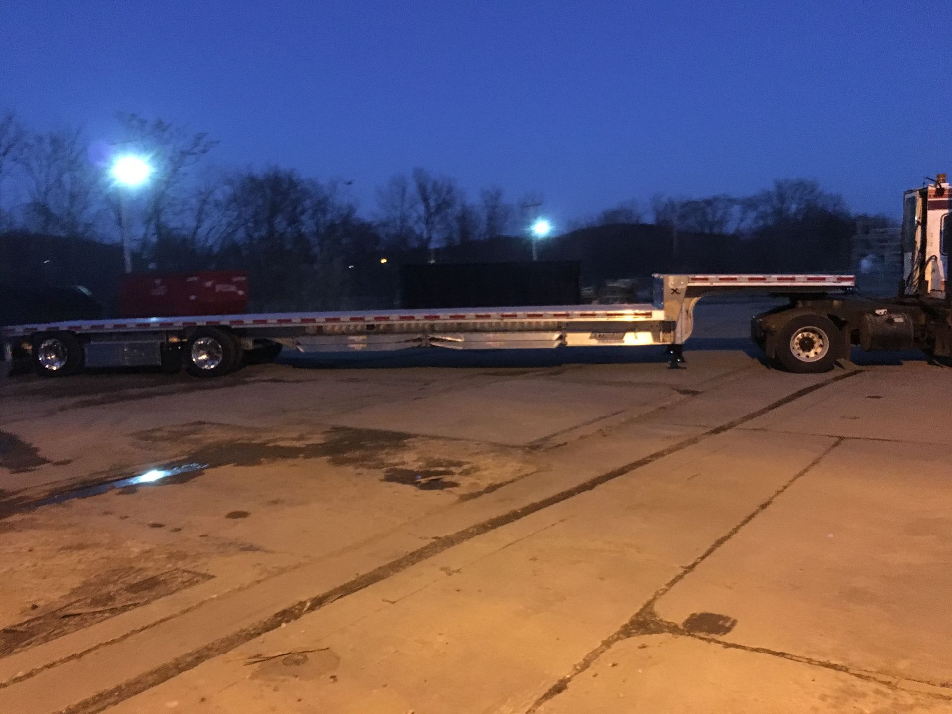Flatbed trailer attached to a semi-truck on a concrete surface at dusk.