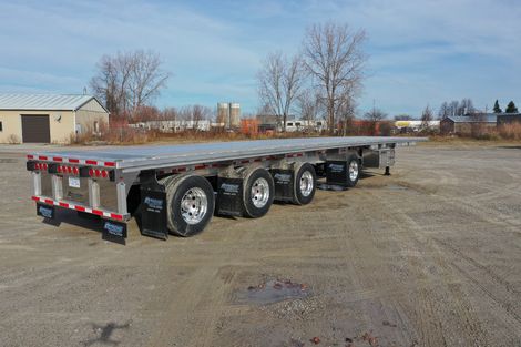 Flatbed semi-trailer parked on gravel. Features include shiny wheels, reflective rear lights, and a gray deck.