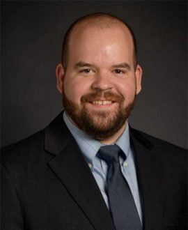 Man in suit smiles at the camera, dark background.
