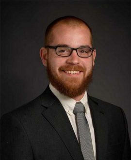 Man in suit and tie, smiling, wearing glasses and a beard, dark background.