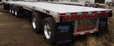 Flatbed trailer with multiple wheels on a dirt surface. Red tail lights and a polished metal finish.