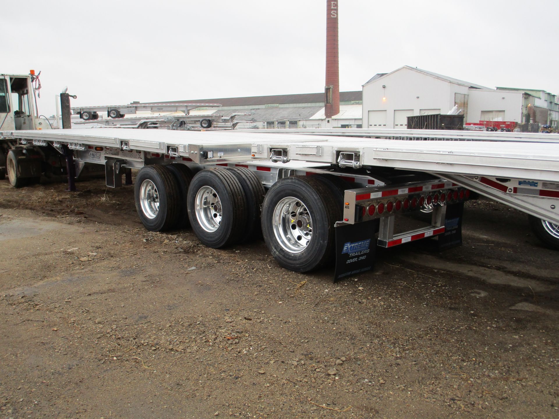 Flatbed semi-trailer with three axles and shiny wheels on a gravel lot; a truck cab is visible.