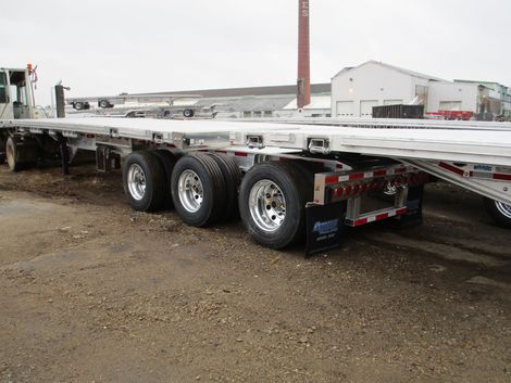 Flatbed semi-trailer with three axles and shiny wheels on a gravel lot; a truck cab is visible.