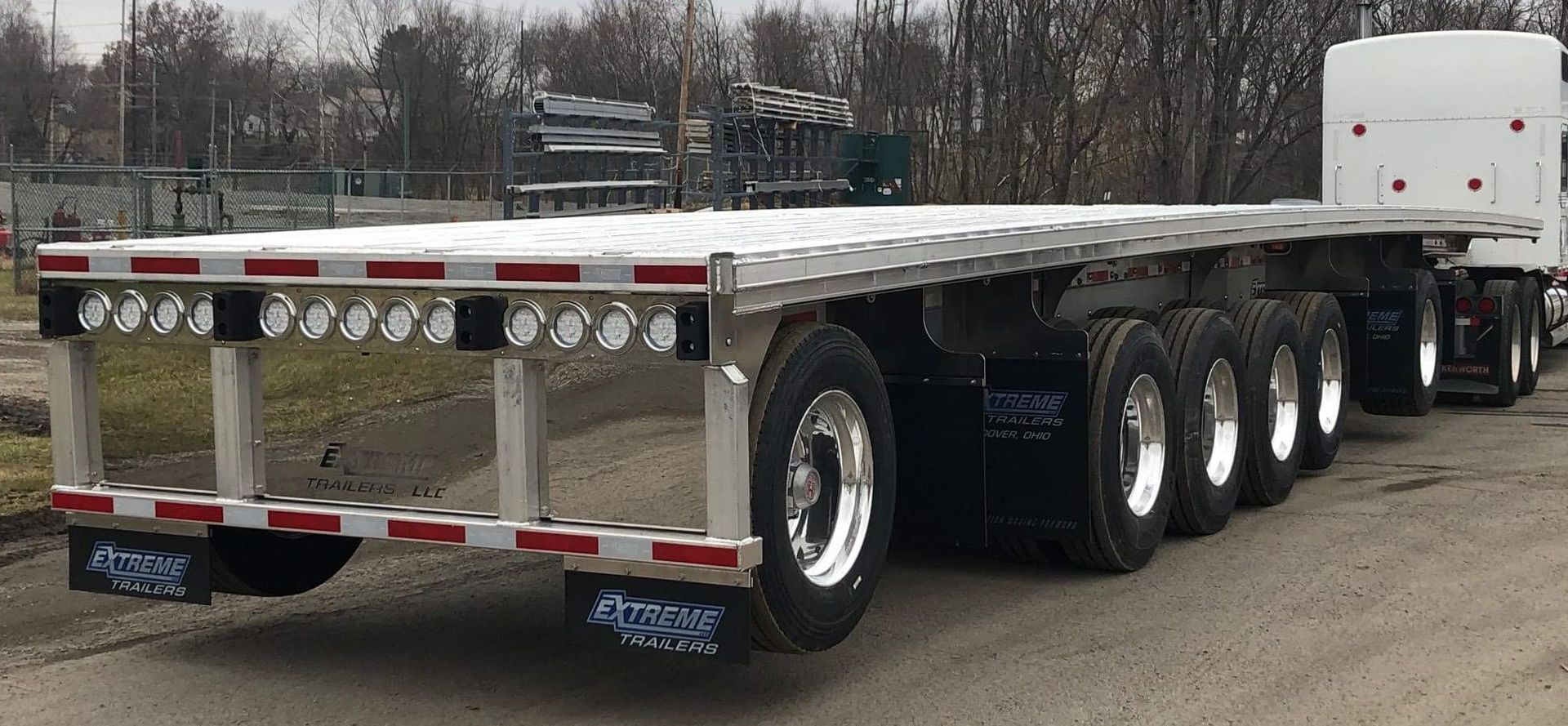 Flatbed semi-trailer parked on pavement; trees in the background. Trailer has many wheels, lights, and reflective tape.