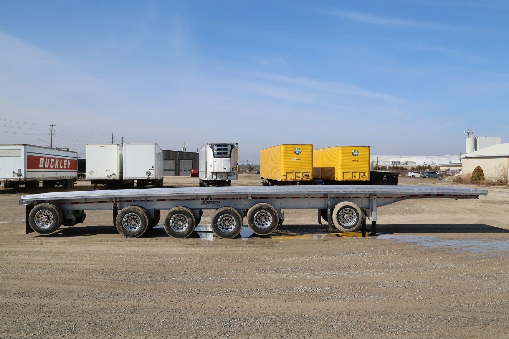 Flatbed trailer with yellow containers, parked outdoors on gravel. Other trailers visible in background.