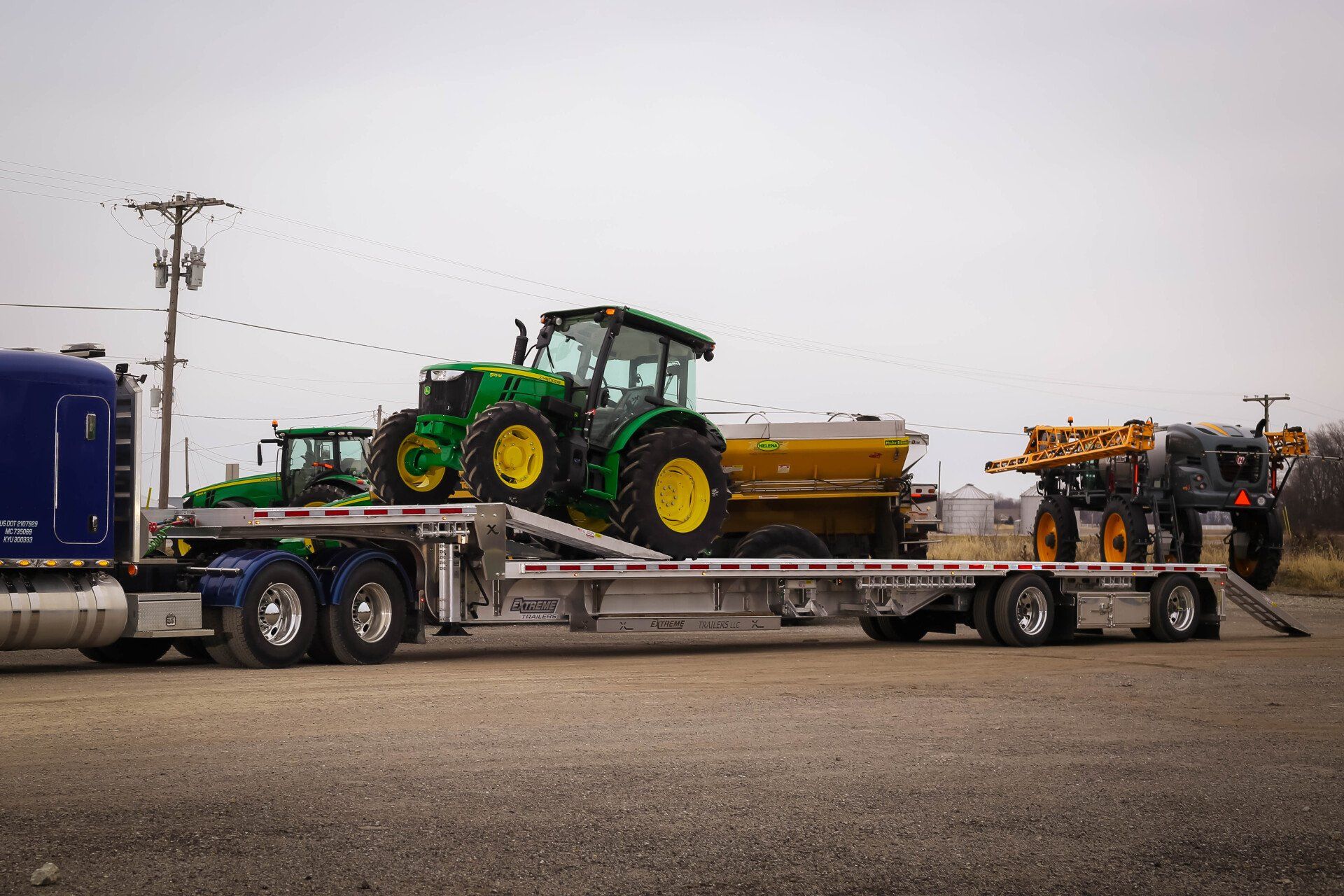A semi-truck hauling three tractors on a flatbed trailer in a rural setting under a cloudy sky.