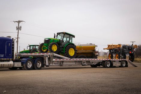A semi-truck hauling three tractors on a flatbed trailer in a rural setting under a cloudy sky.