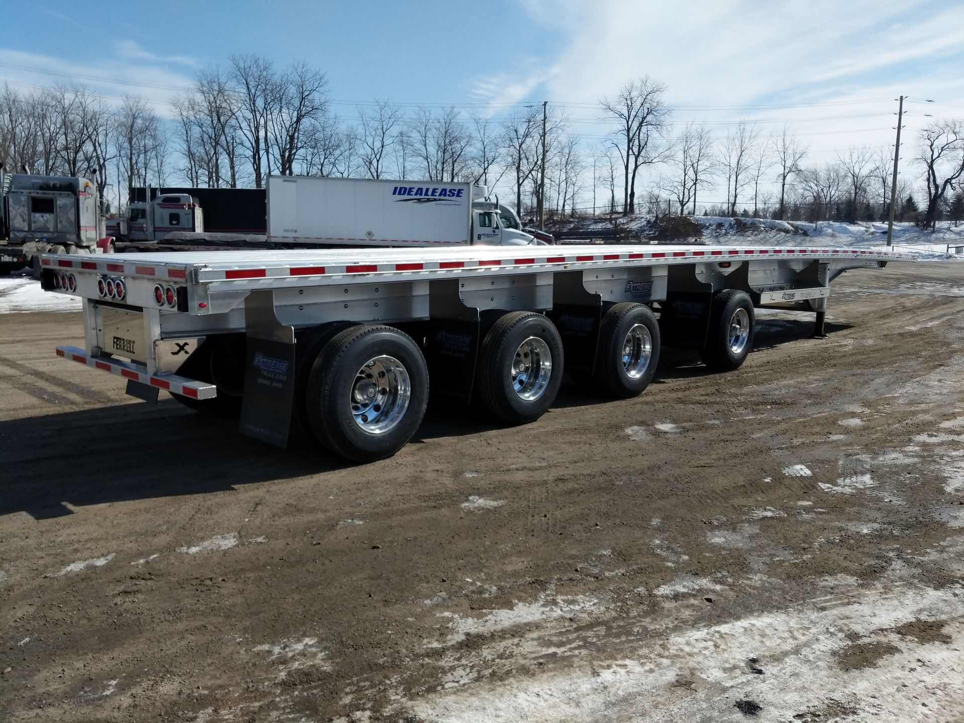 Flatbed trailer on a snow-covered gravel lot. Silver wheels, white deck. Trucks in background. Sunny day.