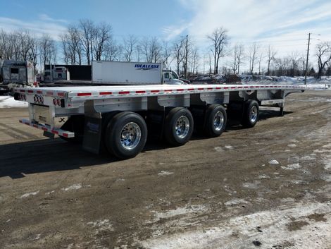 Flatbed trailer on a snow-covered gravel lot. Silver wheels, white deck. Trucks in background. Sunny day.