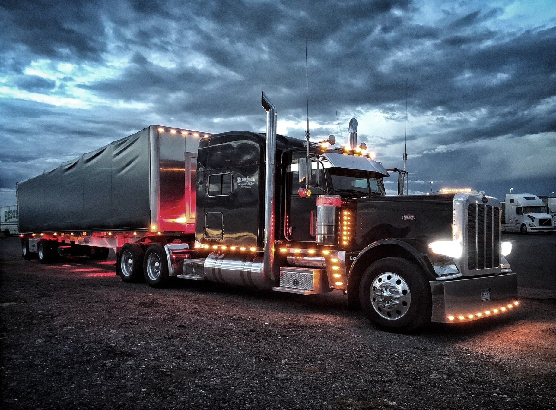 Black semi-truck with trailer, illuminated lights, parked under a cloudy sky.