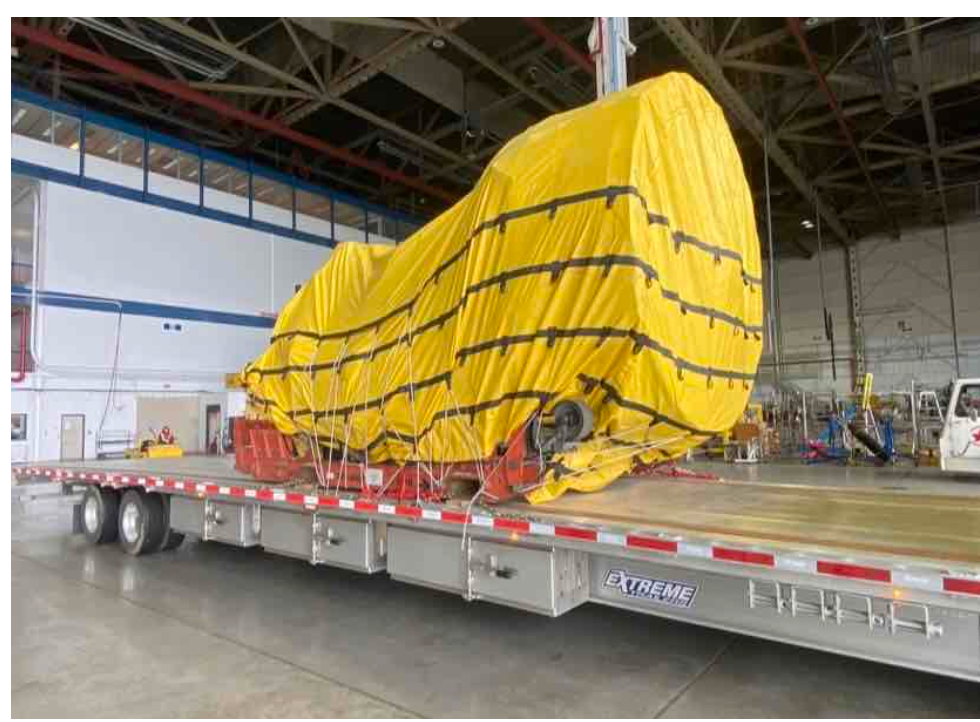 Large, covered machine on a flatbed trailer in a warehouse. Yellow tarp, black straps, red support.