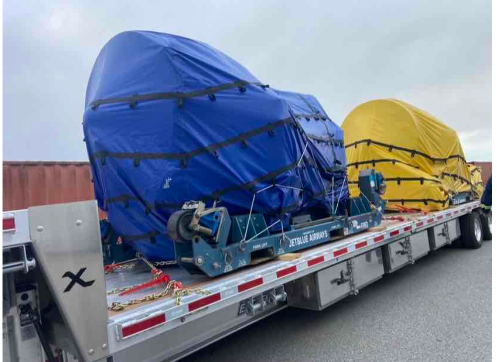 Two large, tarp-covered industrial machines secured to a flatbed truck for transport, one blue and one yellow.