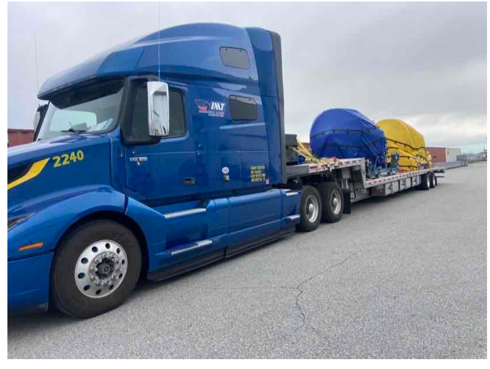 Blue semi-truck pulling a flatbed trailer with large, covered equipment on a paved lot.