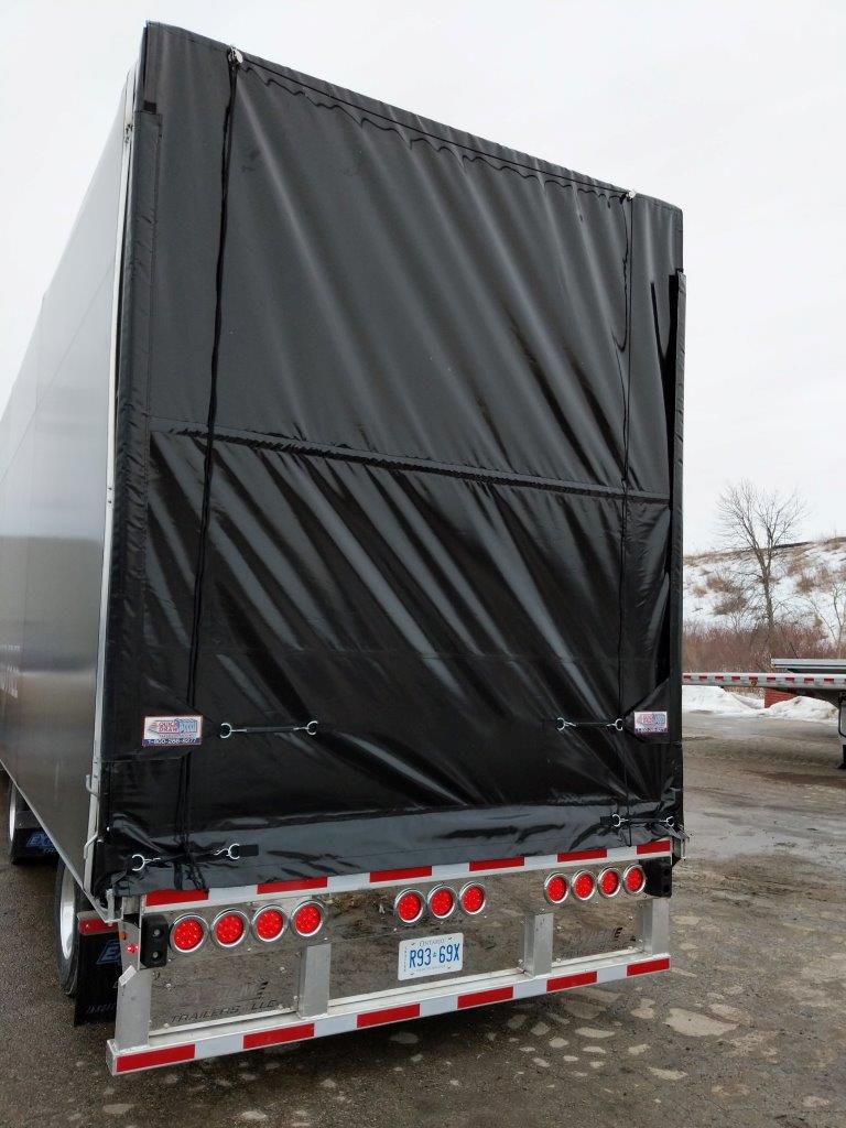 Rear view of a black tarp-covered semi-trailer with red and white taillights in a wintry outdoor setting.