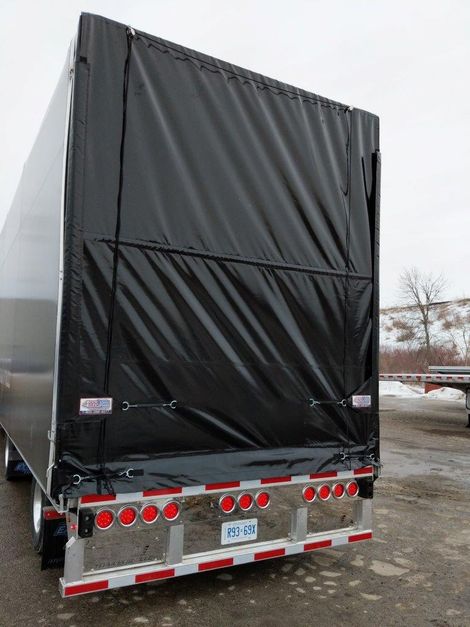 Rear view of a black tarp-covered semi-trailer with red and white taillights in a wintry outdoor setting.