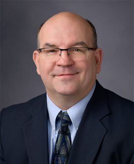 Bald man with glasses wearing a blue shirt, tie, and suit jacket, smiling, studio shot.