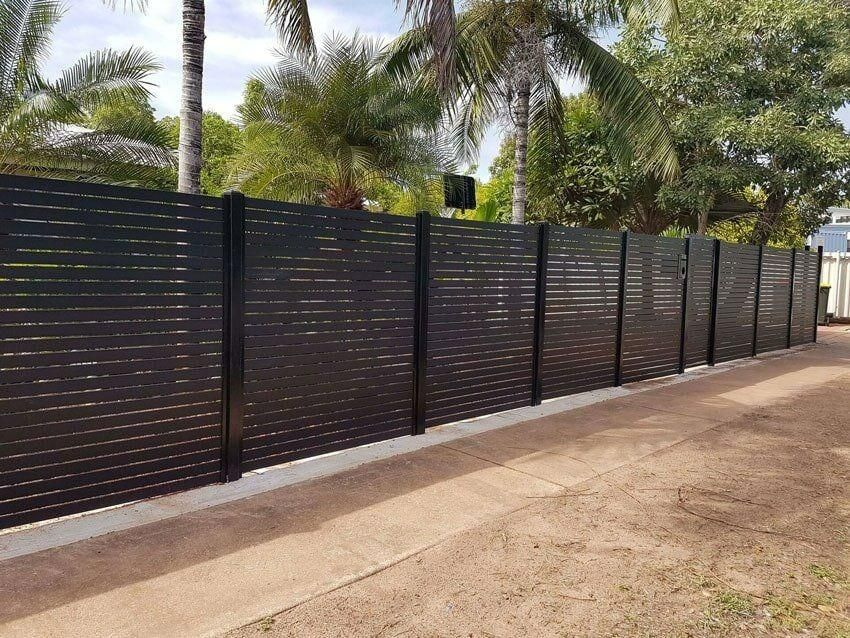 A Black Fence Surrounds A Dirt Road With Palm Trees In The Background — Rabbo's Fencing Services In Pinelands, NT